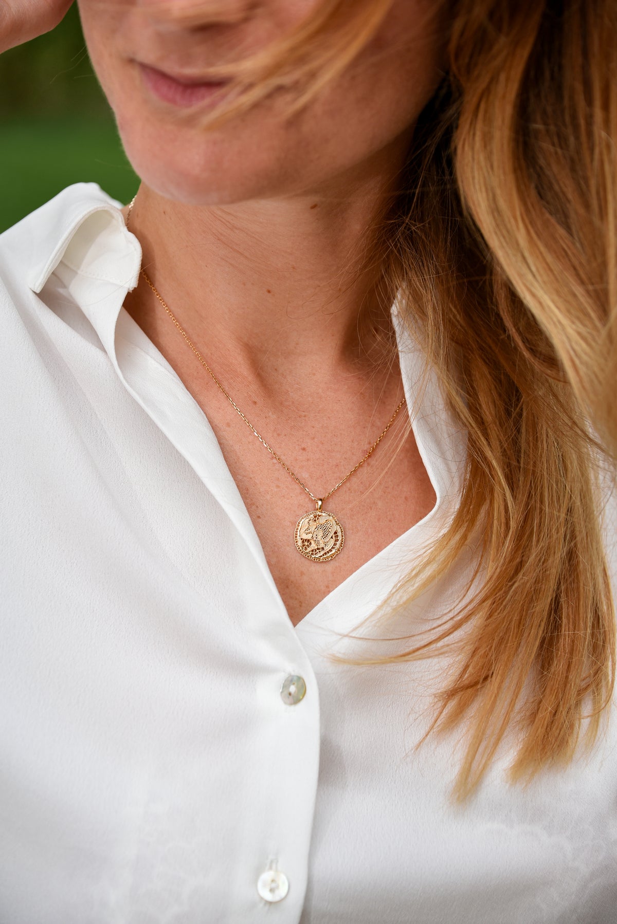 Femme blonde portant le collier Grâce en vermeil doré à l'or 18 carats, avec médaille dentelle ajourée sur une chemise blanche. 