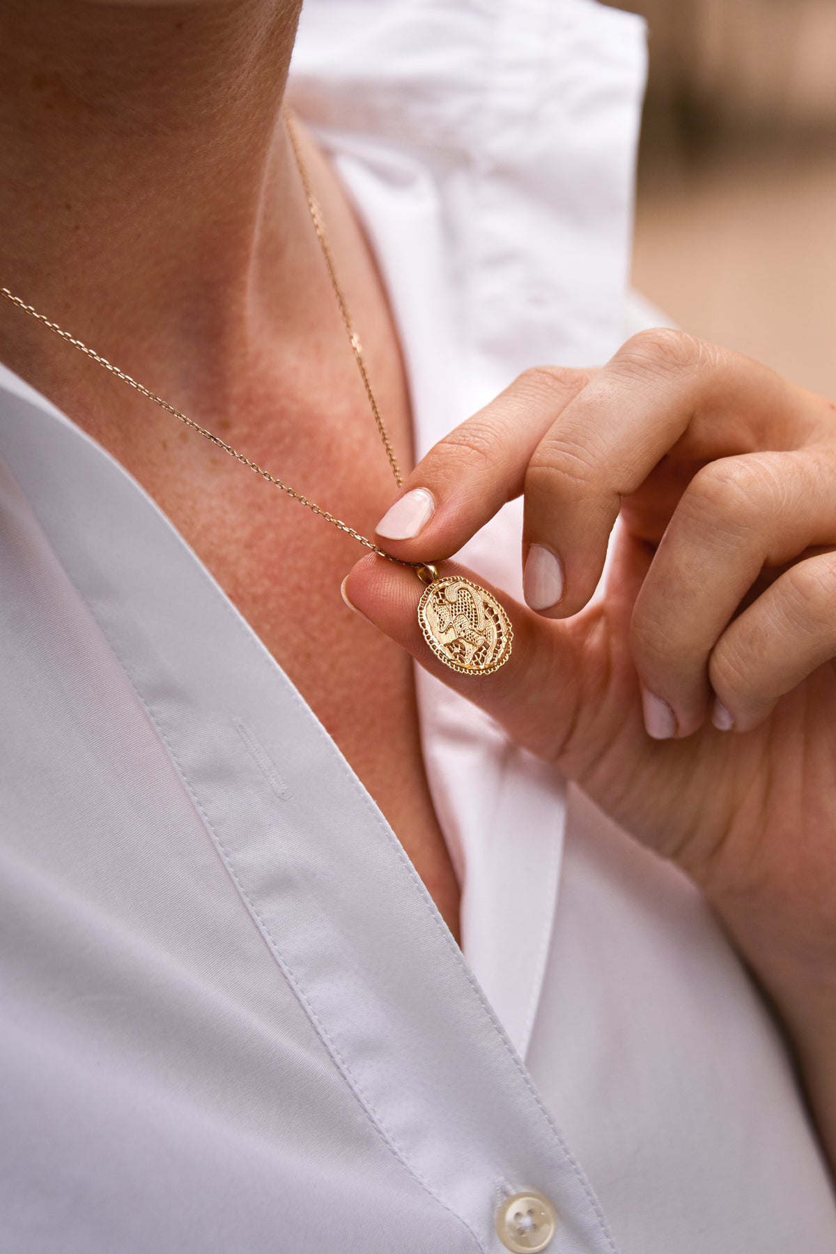 Femme portant le collier Grâce en vermeil doré à l'or 18 carats, tenant délicatement sa médaille dentelle ajourée sur une chemise blanche.
