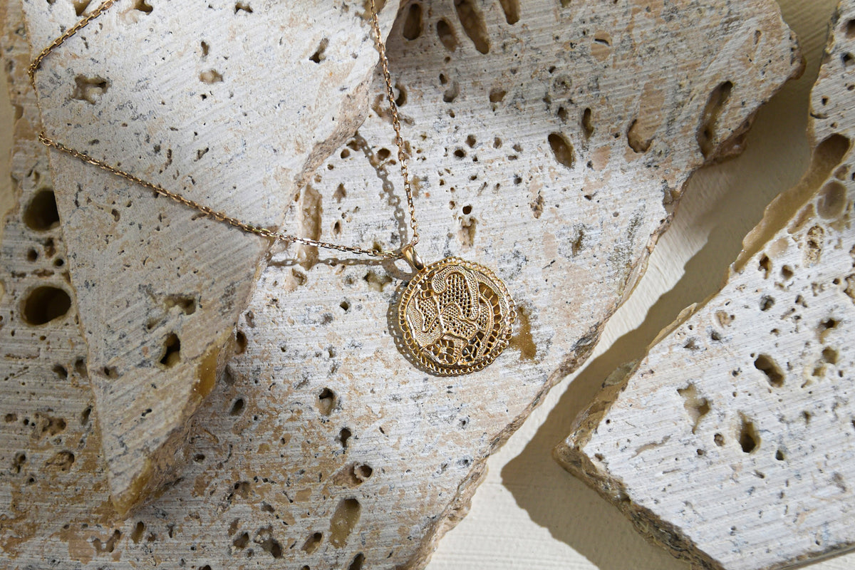 Collier Grâce en vermeil doré à l'or 18 carats, avec médaille dentelle ajourée, délicatement posé sur une pierre en travertin claire.