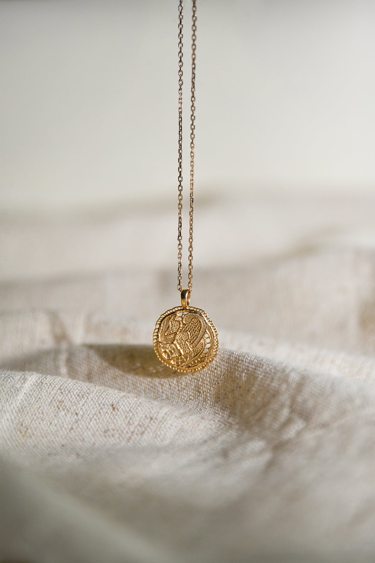Collier Grâce Maison Elise D. en vermeil doré à l'or 18 carats, avec pendentif médaille dentelle ajourée suspendu au-dessus d'un tissu écru en lin.